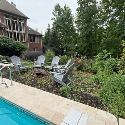 Backyard pool area with Adirondack chairs, greenery, and a fire pit beside a brick house.