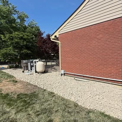 Gravel pathway beside a brick house with a pool equipment area and green trees.